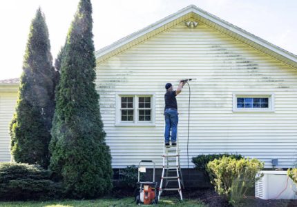Real life, real person DIY senior man is balanced near the top of a ladder while cleaning the vinyl clapboard siding on his house with high pressure cleaning power wash equipment. Very satisfying to see such clear progress as he methodically moves the spray nozzle back and forth across the grungy grimy surface!